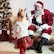 A young girl is walking toward a smiling seated Santa Claus in front of a Christmas tree.
