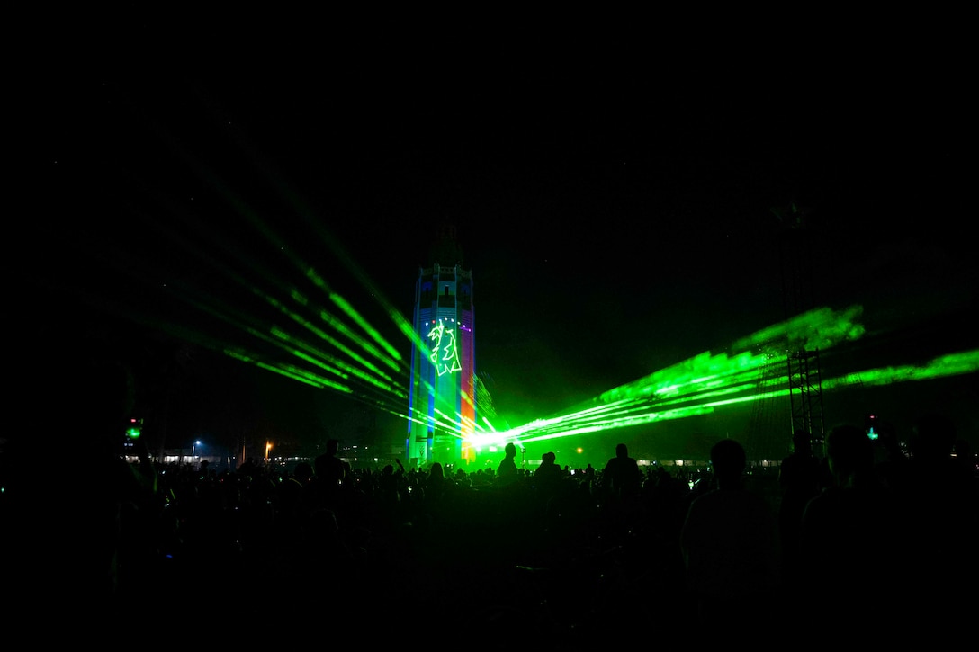 Streaks of green light shine from a tower over a crowd at night.
