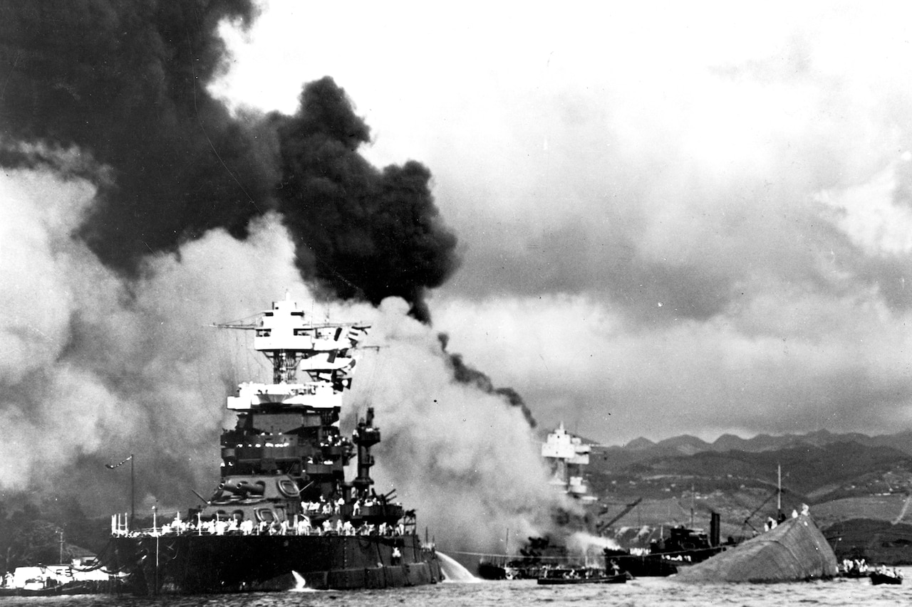 A ship sits in water near a capsized ship as smoke billows.
