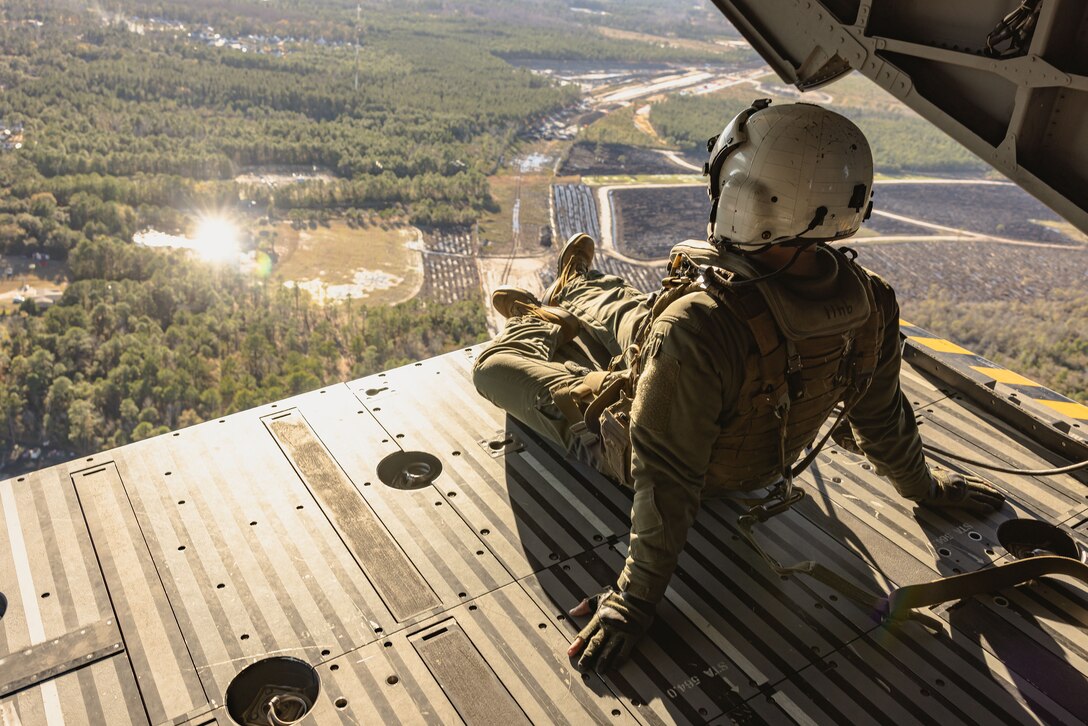 A uniformed Marine wearing a helmet views the terrain below while sitting inside an open military aircraft.