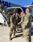 Soldiers in uniform walking down an airplane stairs ramp. carrying bags, and shaking hands with Soldier leadership. It's during the day with bright sunshine and a bright blue sky. The soldiers are various races, ages and genders.