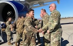 Soldiers in uniform walking down an airplane stairs ramp. carrying bags, and shaking hands with Soldier leadership. It's during the day with bright sunshine and a bright blue sky. The soldiers are various races, ages and genders.