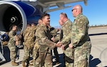 Soldiers in uniform walking down an airplane stairs ramp. carrying bags, and shaking hands with Soldier leadership. It's during the day with bright sunshine and a bright blue sky. The soldiers are various races, ages and genders.