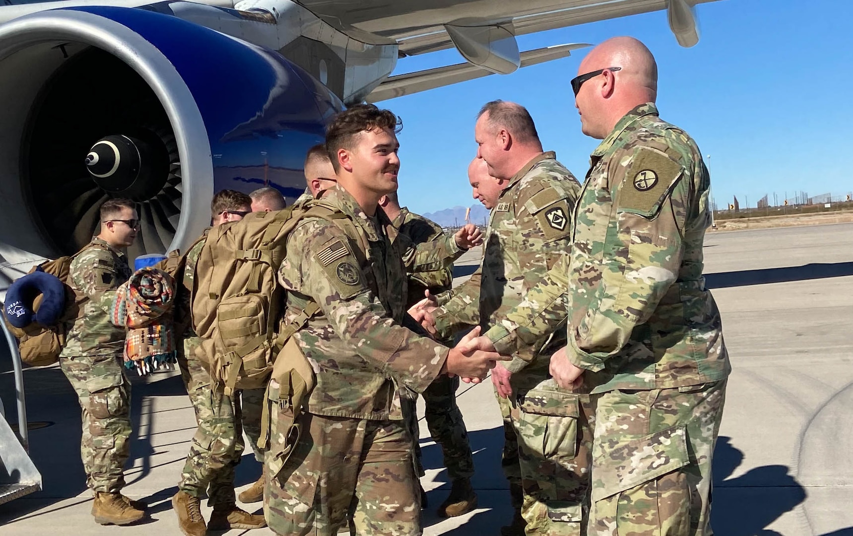 Soldiers in uniform walking down an airplane stairs ramp. carrying bags, and shaking hands with Soldier leadership. It's during the day with bright sunshine and a bright blue sky. The soldiers are various races, ages and genders.