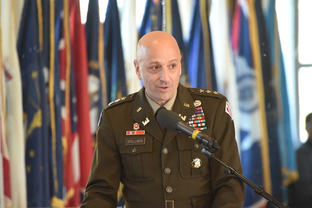 A man in Army uniform speaks at a podium. Flags appear in the background.