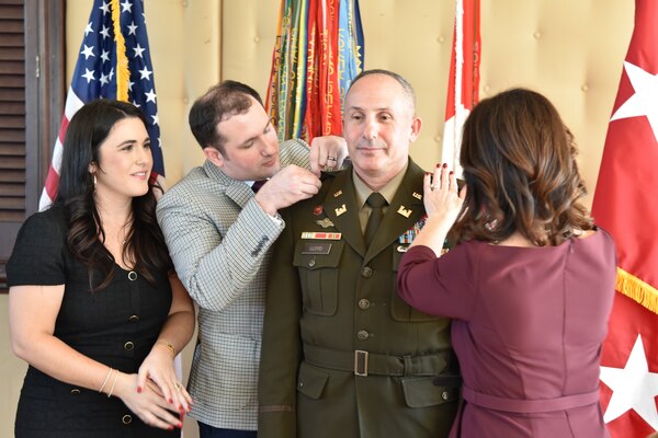 A man and woman pink Army rank on the uniform of an Army officer while another woman looks on.