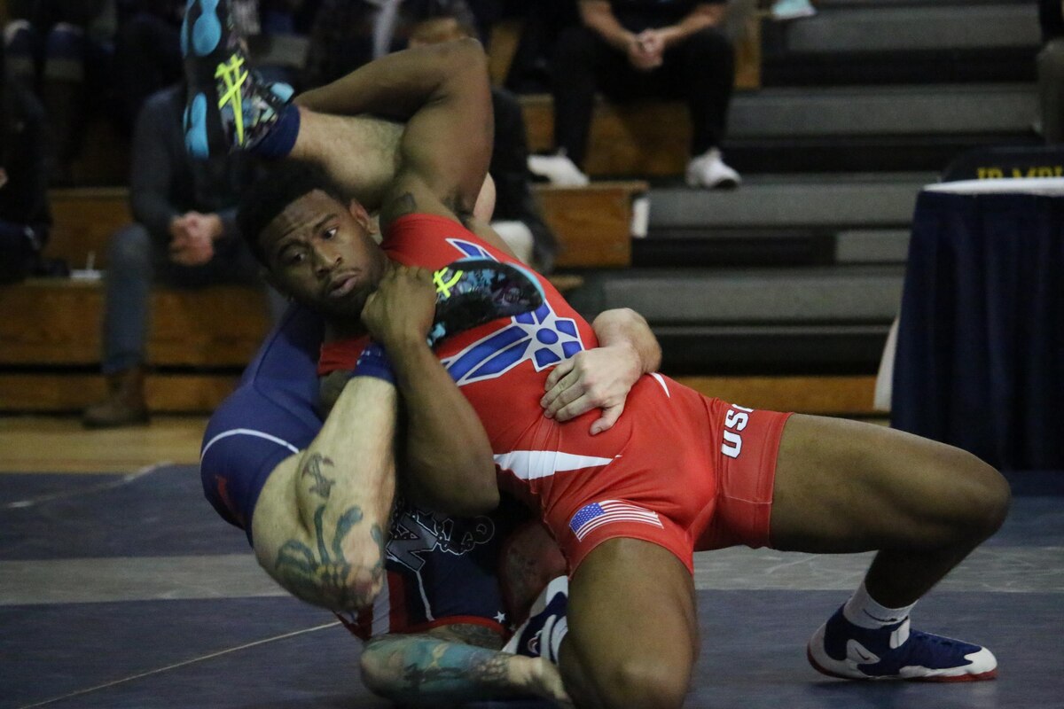 First athlete on his knee with second athlete at his back, with shoulders on the ground and one arm around first athlete's waist, trying to wrap his legs around the first athlete. First athlete tries to secure second athletes feet and legs.