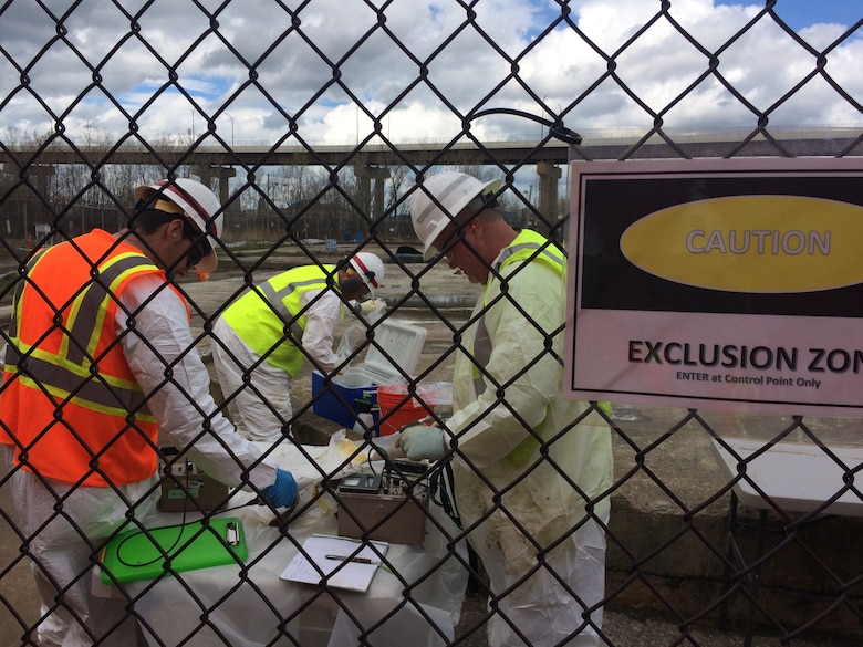 Three men in white coveralls, reflective vests, and hard hats use diagnostic equipment to conduct groundwater sampling behind a black chain-link fence with a caution sign.