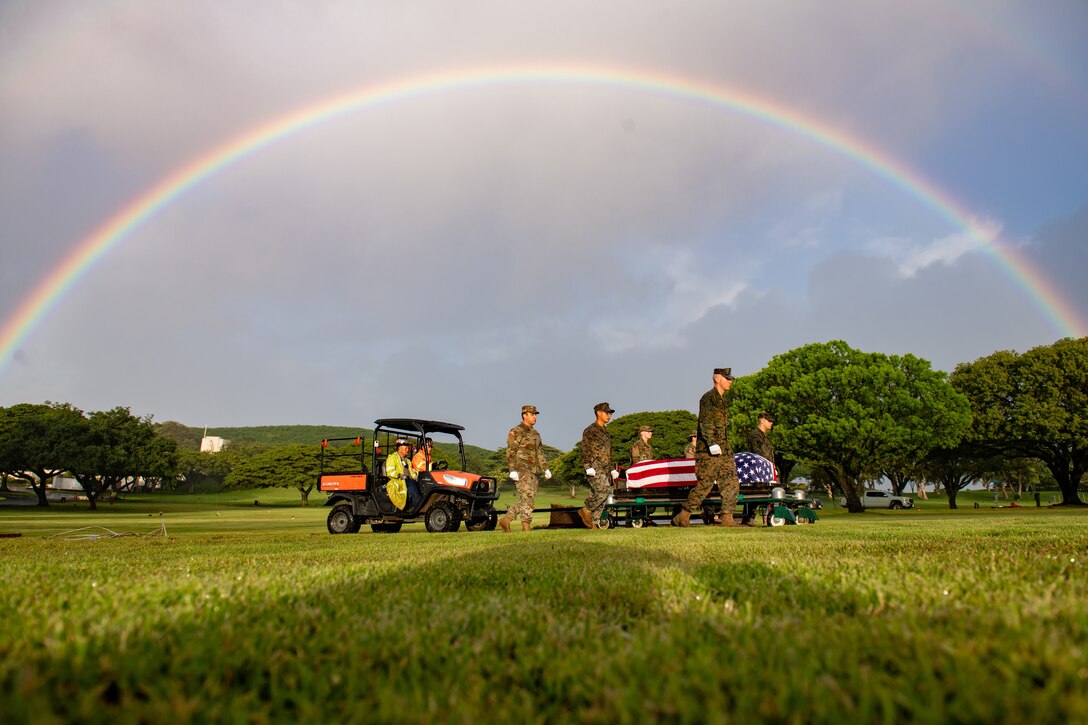 Service members walk alongside a casket while a small vehicle follows. A rainbow fills the sky behind them.
