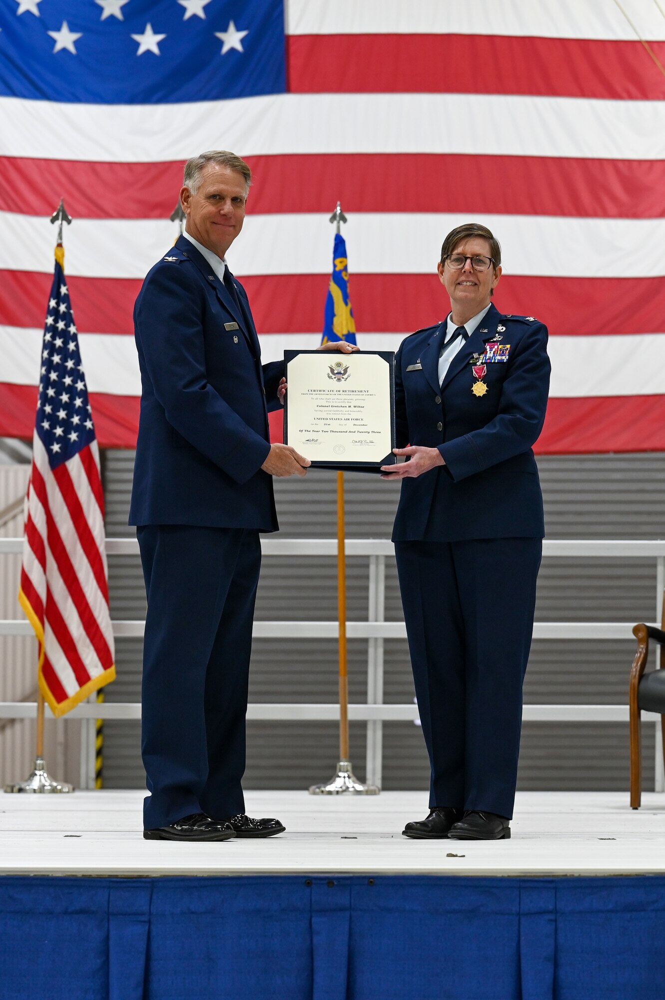 Larry Shaw and Gretchen Wiltse, both in Air Force dress uniforms, pose with a retirement certificate on a stage directly in front of a large American flag.