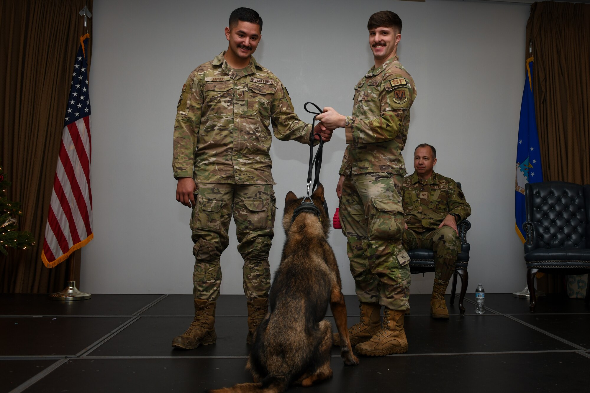 Two men in military uniforms stand on stage with a dog while holding onto its leash.
