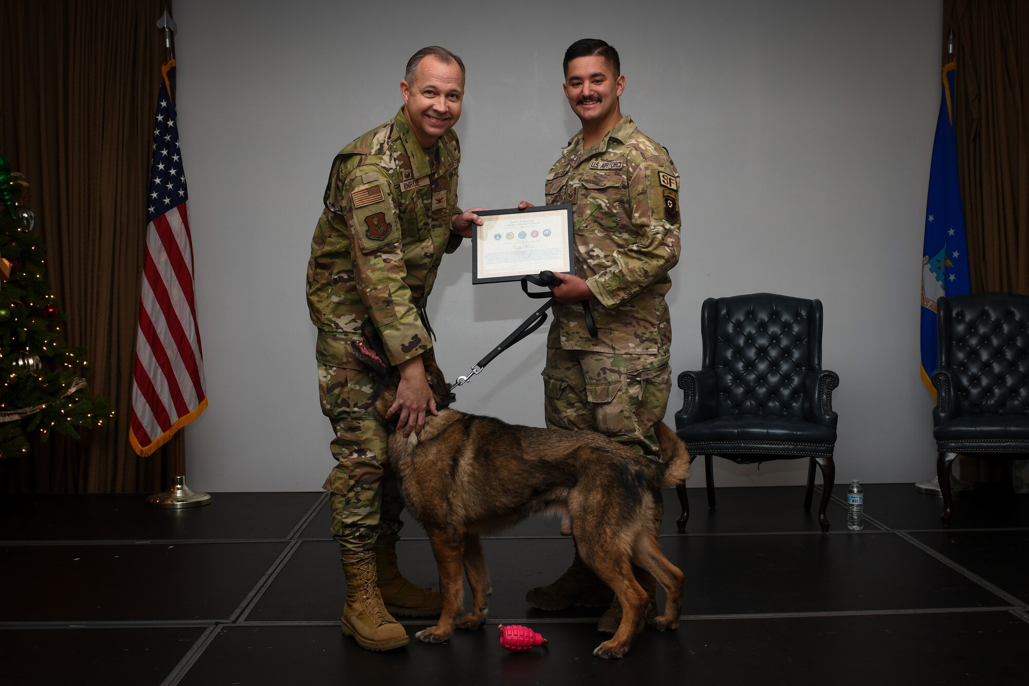 Two men in military uniforms stand on stage with a dog while holding a certificate.