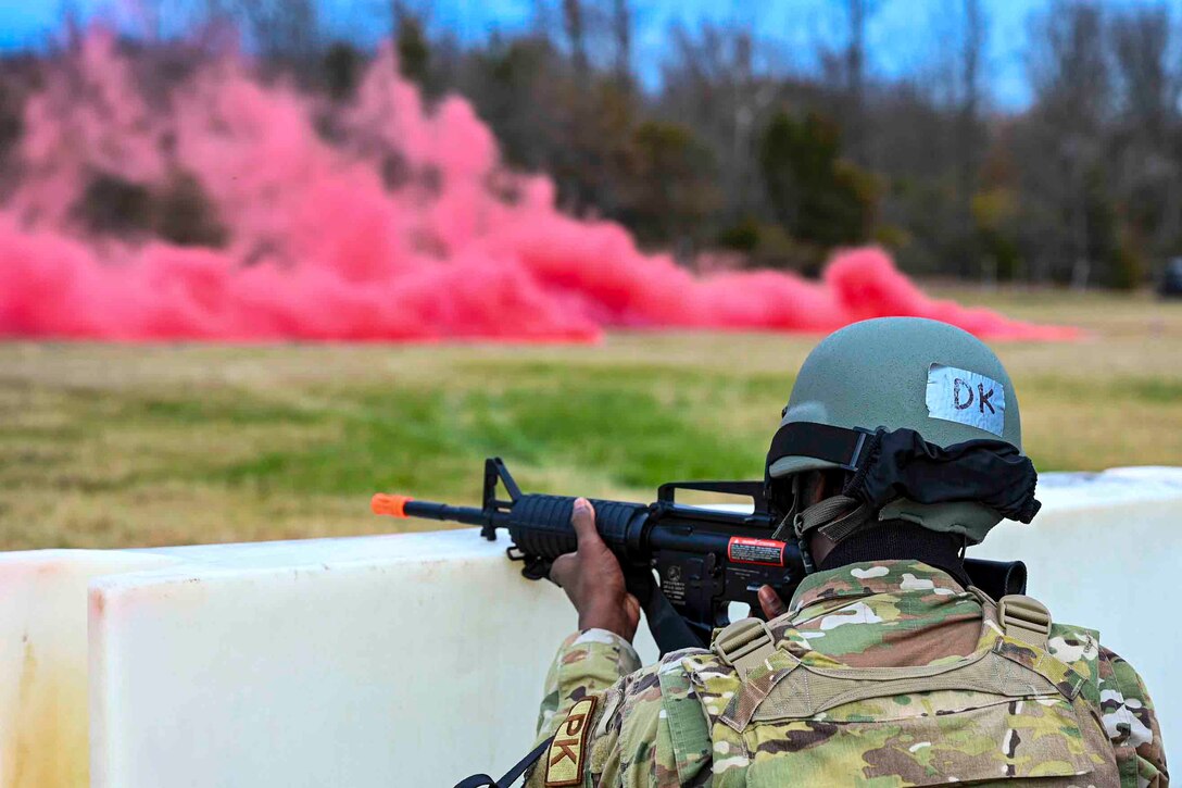 An airman points a weapon from behind a barrier toward a field with pink smoke rising from the ground.
