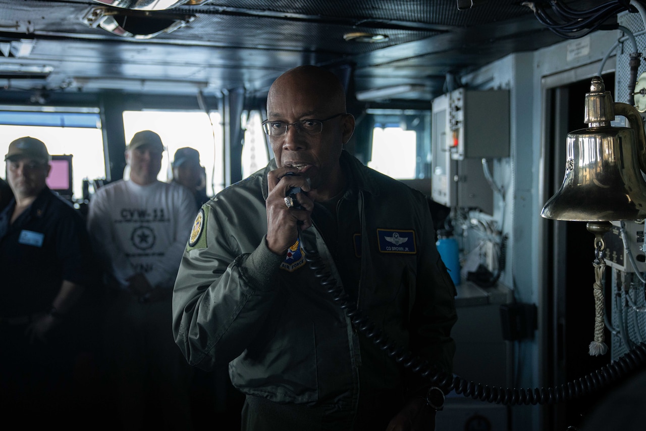 A person in uniform speaks into a microphone from the bridge of a ship.