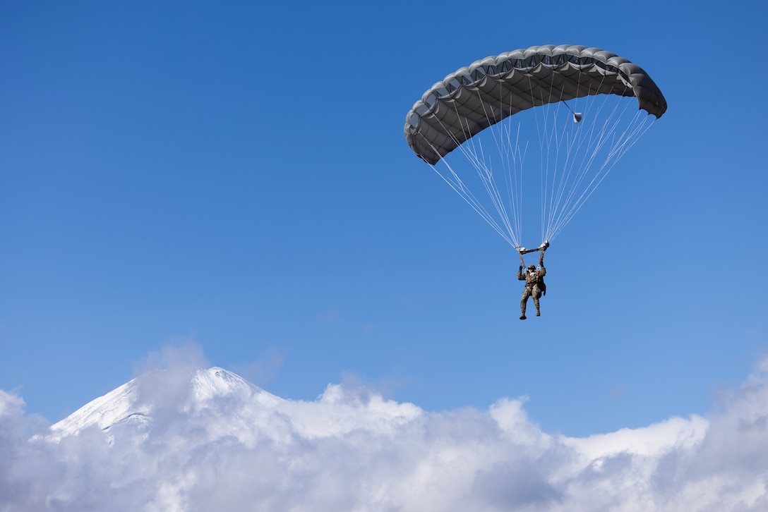 A Marine wearing a parachute descends toward the ground during daylight. A snow-capped mountain and clouds can be seen in the distance.