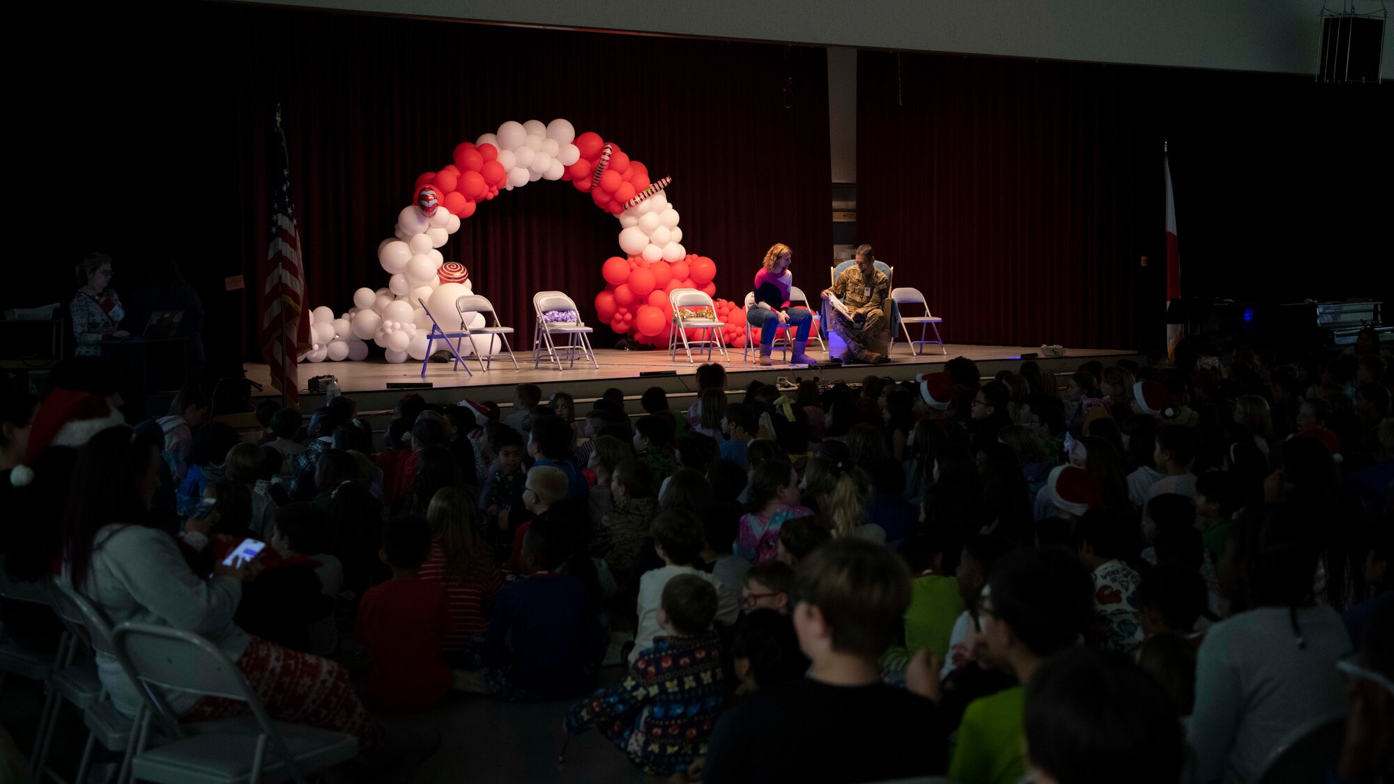 A group of students look towards a stage where a military man and a woman sit reading a book.