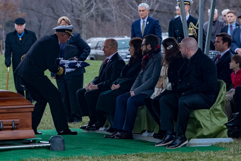 A person hands a folded flag to a man in a row of seated people at a burial.