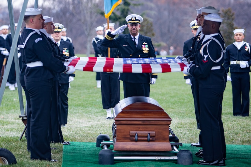 A person salutes a U.S. flag being held over a casket by six sailors.