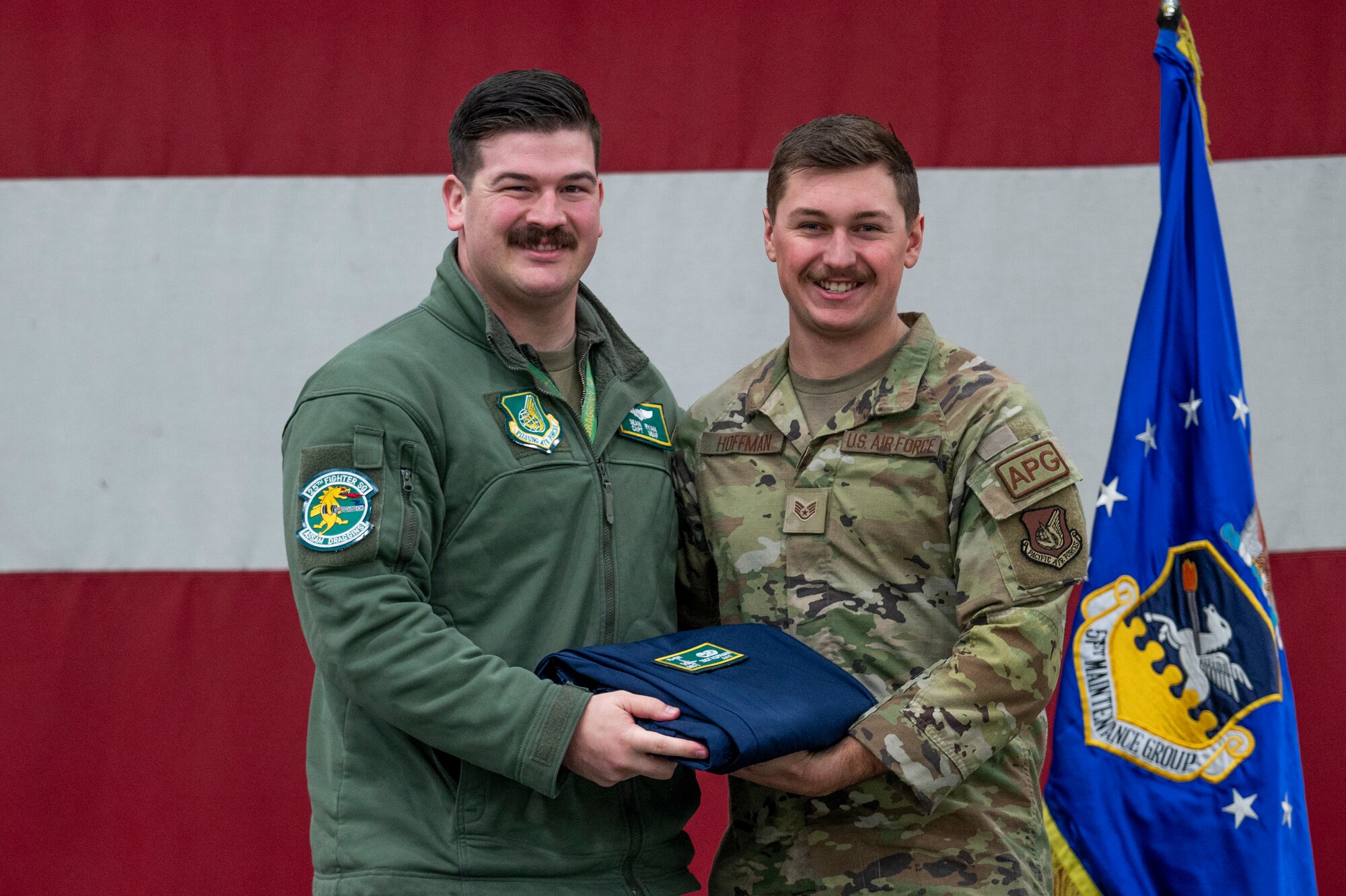 U.S. Air Force Capt. Sean Ryan, 25th Fighter Squadron pilot, presents Staff Sgt. Connor Hoffman, 25th Fighter Generation Squadron, with a patch and coveralls during the 25th FGS Dedicated Crew Chief Ceremony at Osan Air Base, Republic of Korea, Dec. 19, 2023.