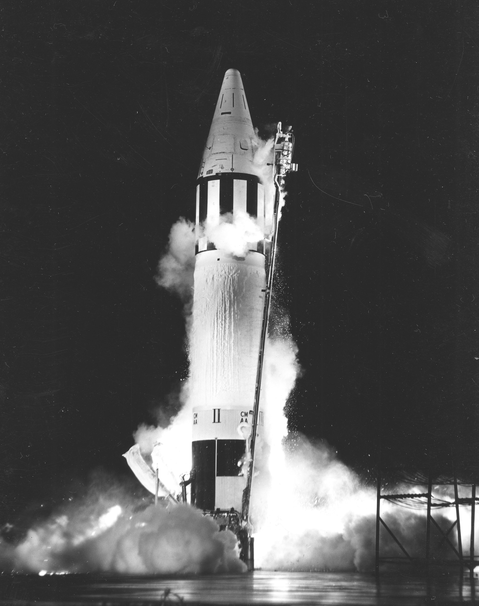 Black and white image of the Jupiter ballistic missile, white with black stripes across the top, on a black background with fire and smoke billowing up from the bottom as it takes off.