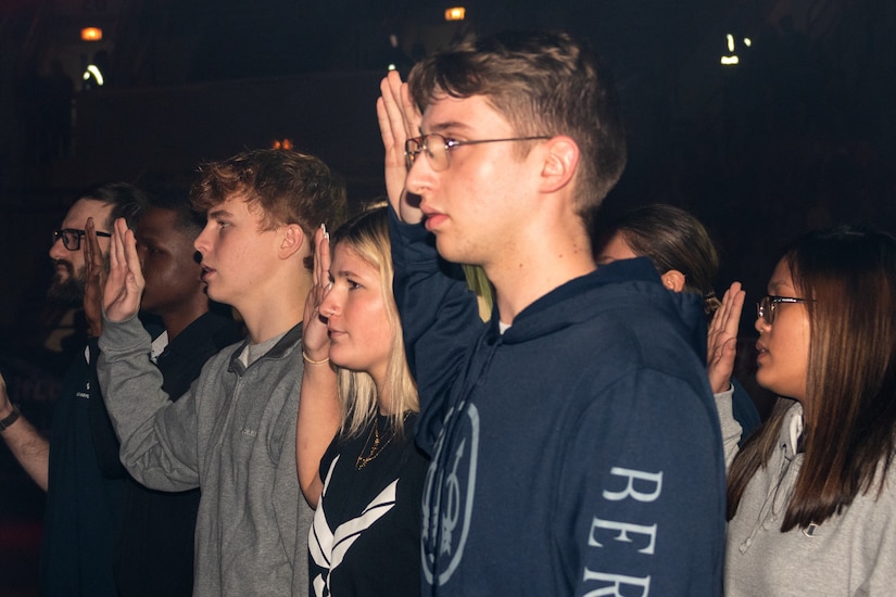 Young women and men, standing in a group, raise their right hands.