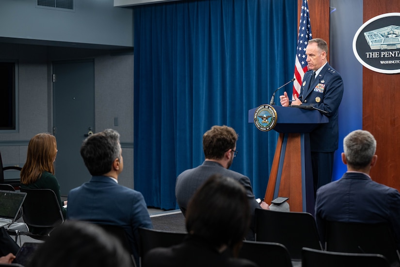 A man in a uniform stands at a lectern and addresses a group of people who are seated in front of him.