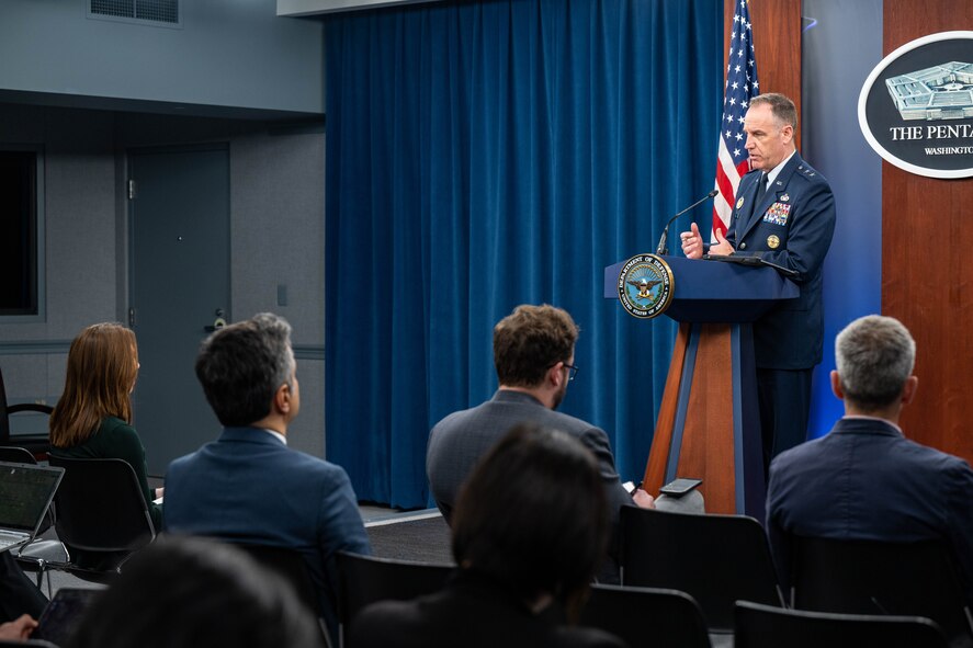 A man in a uniform stands at a lectern and addresses a group of people who are seated in front of him.