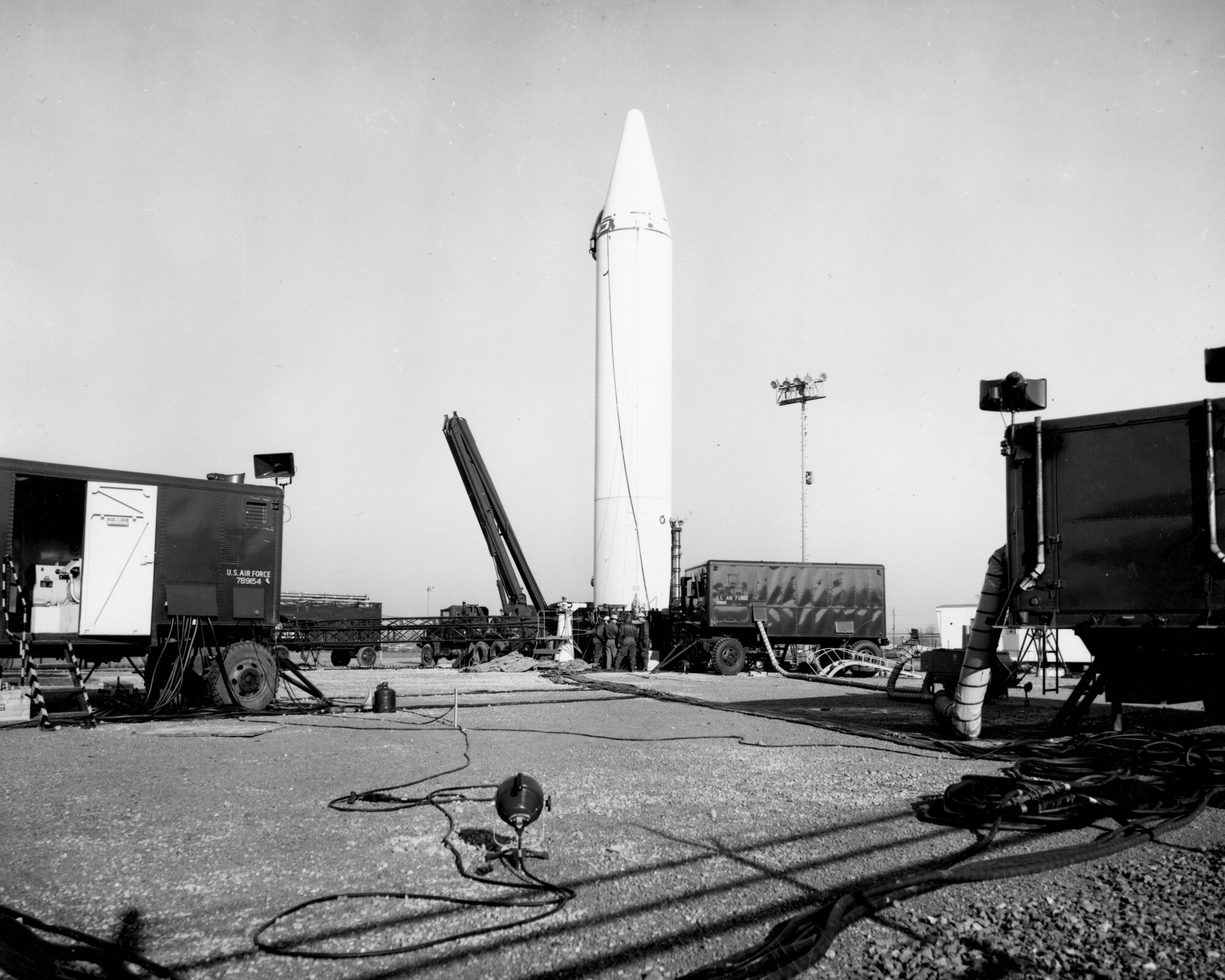Jupiter ground support equipment was complex, involving vehicles carrying liquid oxygen, hydraulic and pneumatic equipment, generators, and other tools.
Black and white image of the white jupiter missile surrounded by servicing equipment.