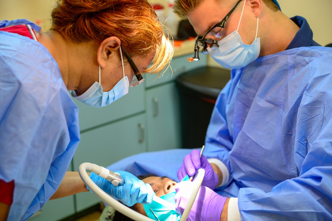 Two medical professionals in blue gowns work on a patient's mouth.