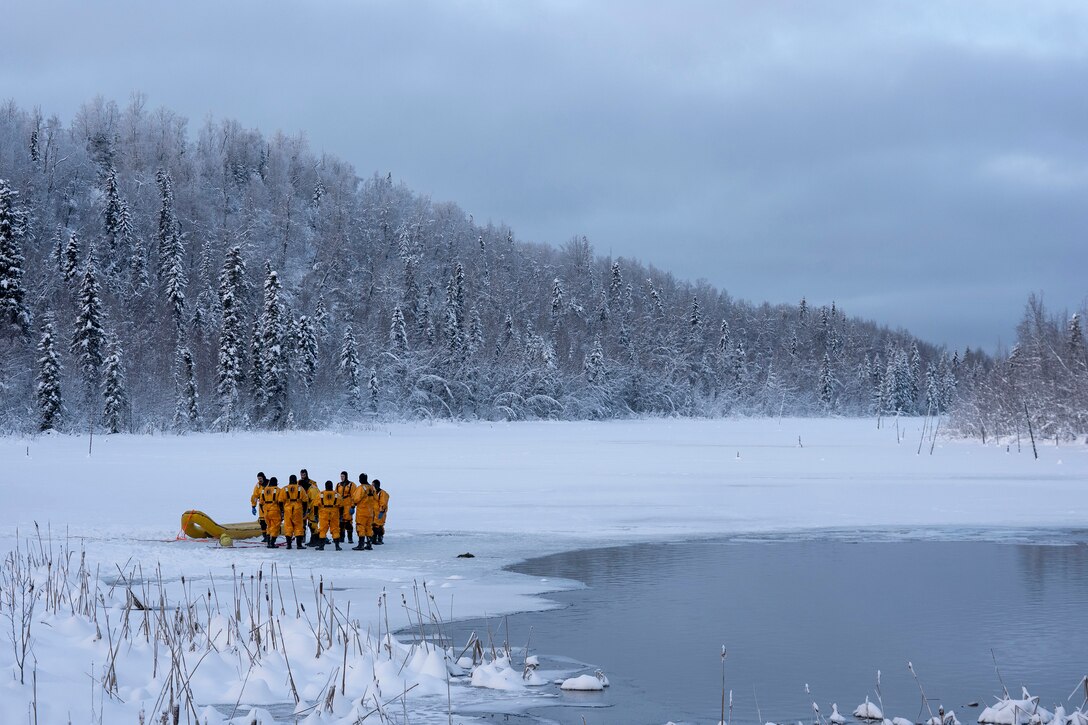 People in yellow protective gear stand by a yellow inflatable raft on a snowfield by a lake.