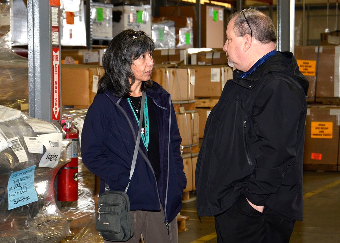 Kathy and Arthur stand facing each other talking inside a warehouse.