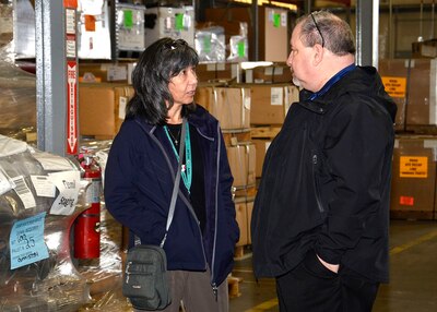 Kathy and Arthur stand facing each other talking inside a warehouse.
