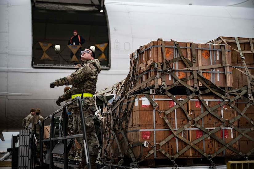 A service member in uniform stands next to pallets of military cargo.