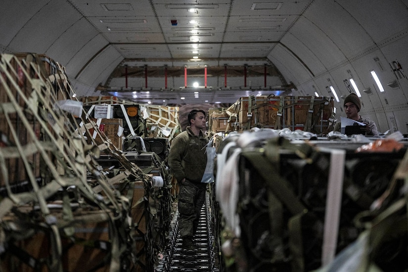 Service members in uniform survey military cargo loaded onto a plane.