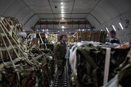 Service members in uniform survey military cargo loaded onto a plane.