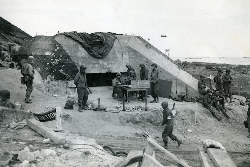 Service members wander around a makeshift command post set up on the bluff of a beach beside a pillbox.