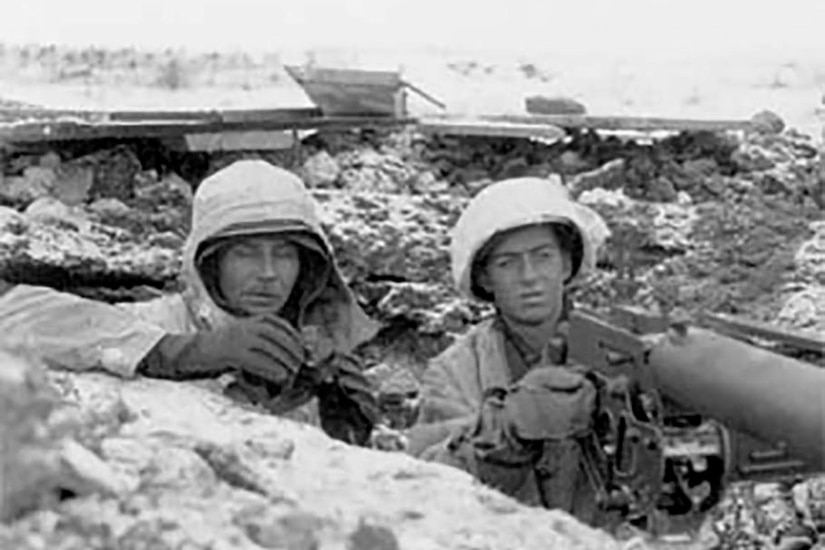 Two helmet-clad men in a chest-deep ditch hold pairs of binoculars.