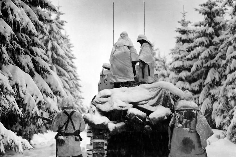 Two people stand on a snow-covered vehicle while two others walk behind it. Trees are covered in snow.