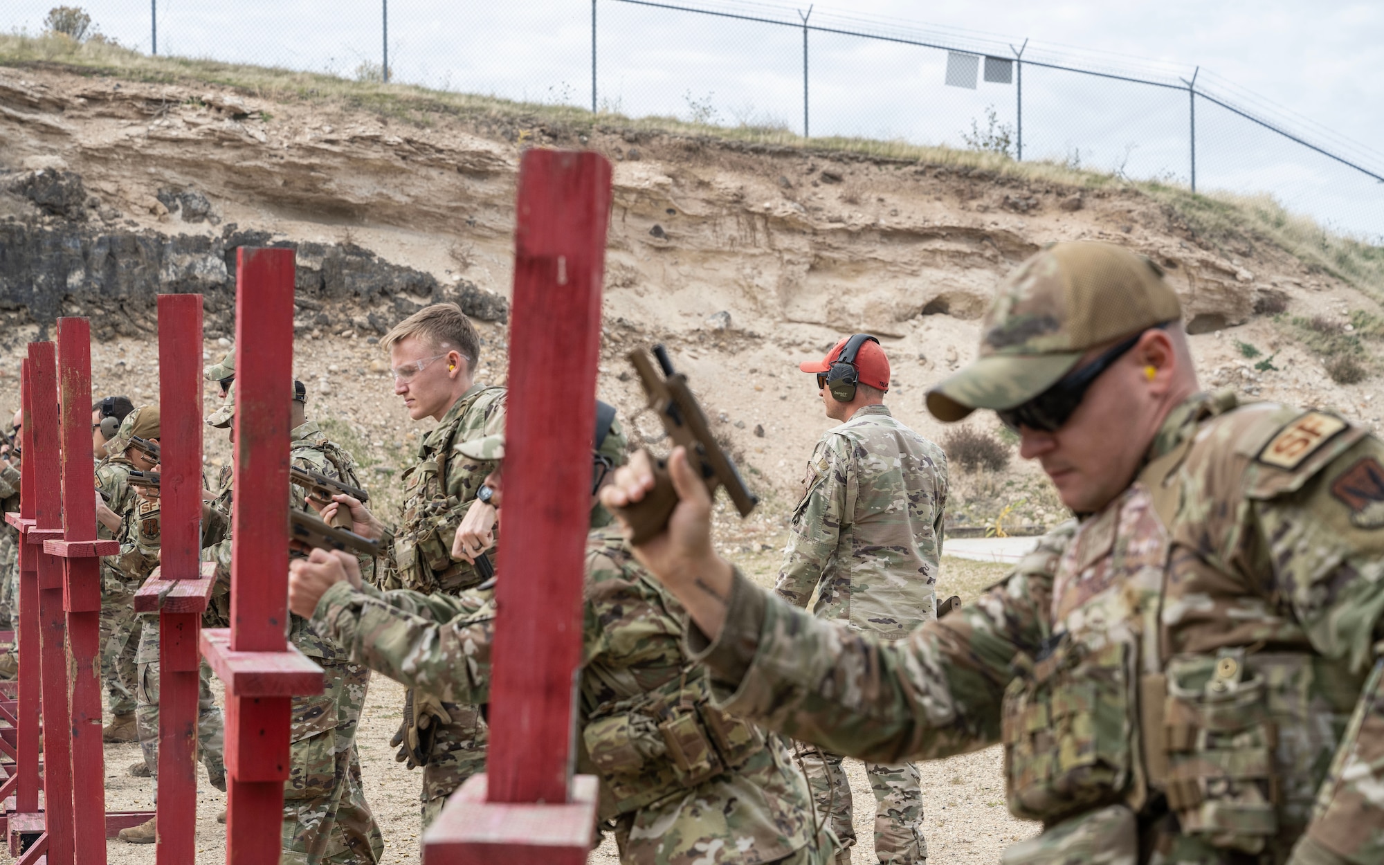 Multiple United States Air Force Security Forces Airmen stand behind individual mock barriers and shoot their side arms while an instructor wearing a red hat observes.