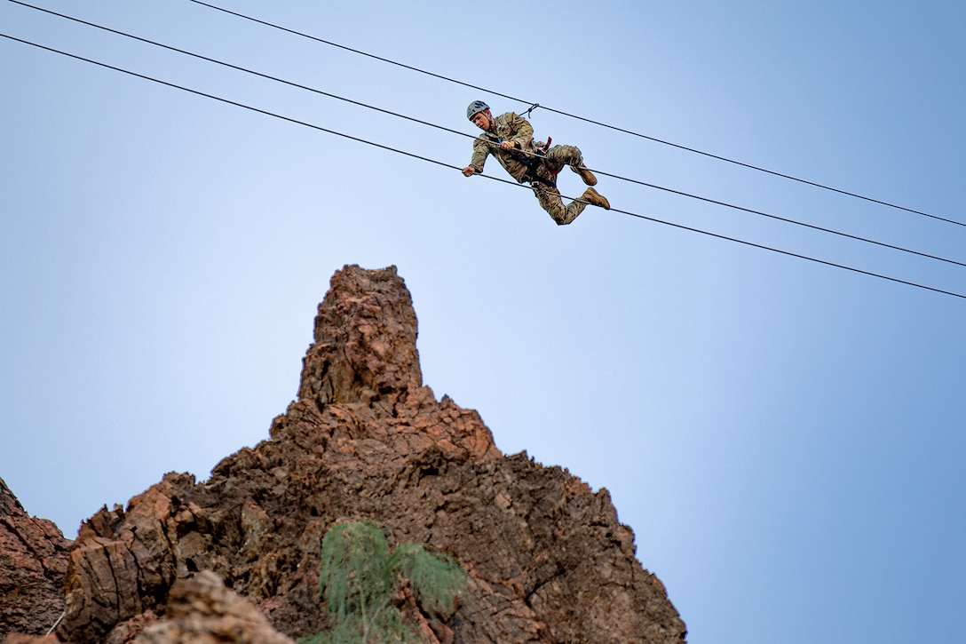 A uniformed solider wearing a helmet maneuvers through high wires with a rock formation visible in the background.