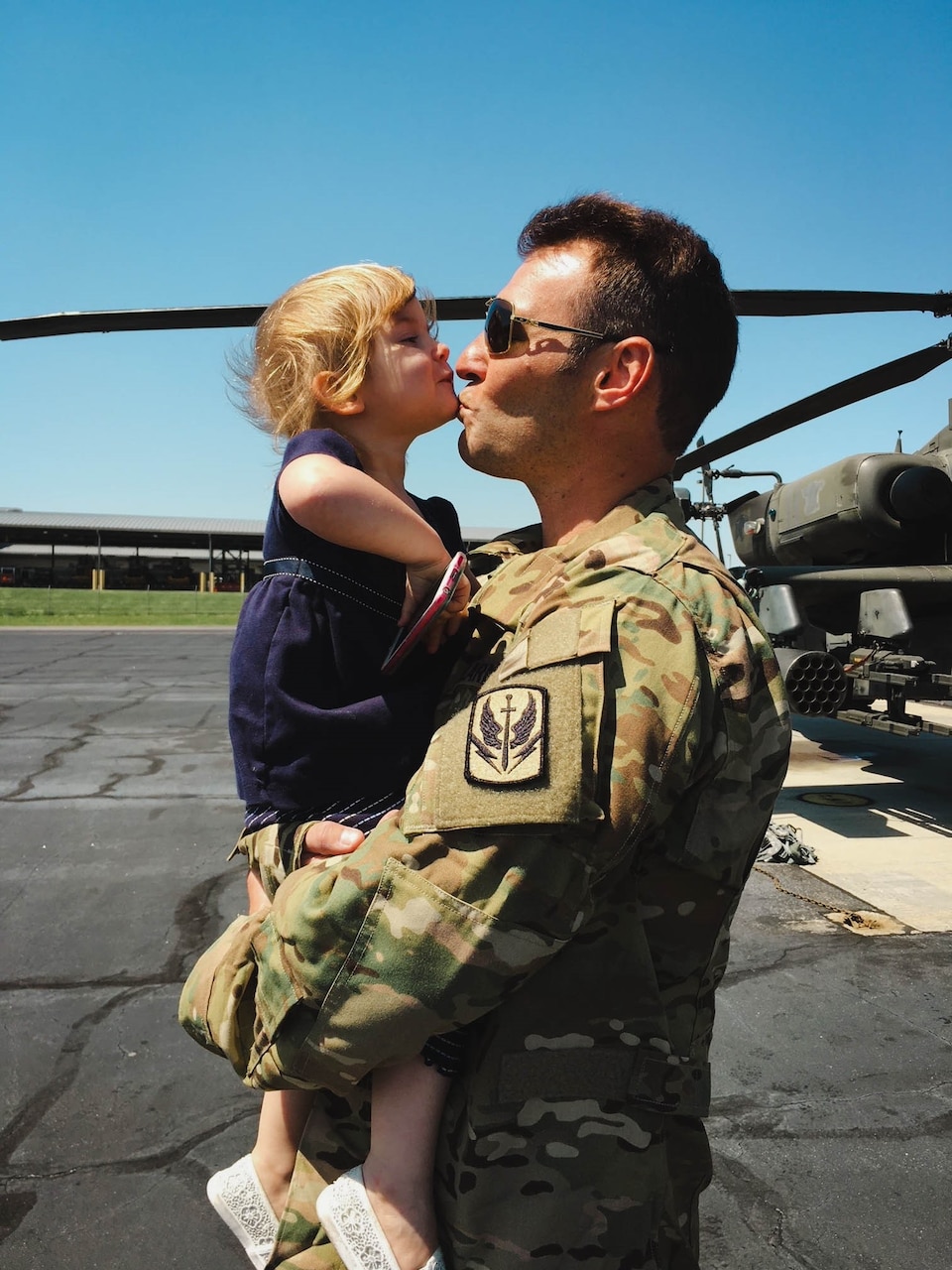 An airman holds a small child as they kiss each other on a flight line, with a helicopter in the background.