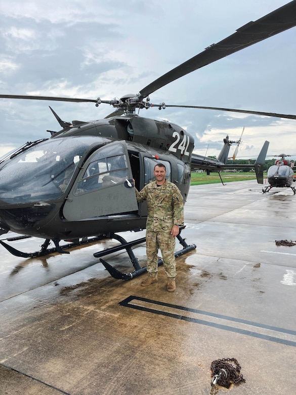 An airman stands in front of a parked helicopter on a flight line.