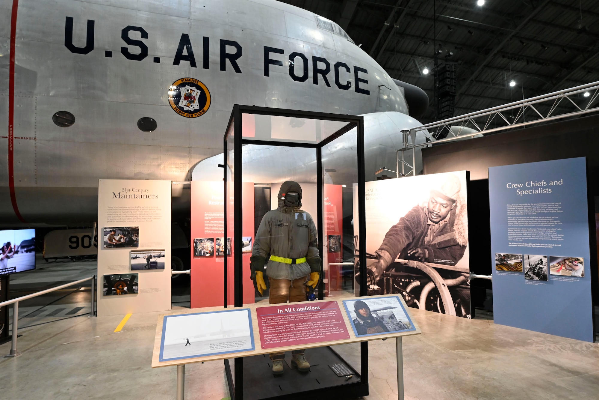 Image of the Maintainer portion of the enlisted exhibit. In the front is a vertical clear case with a cold weather work uniform in it and behind it is a series of vertical panels with images of maintainers on them. Behind it is the nose of the C-124 aircraft.