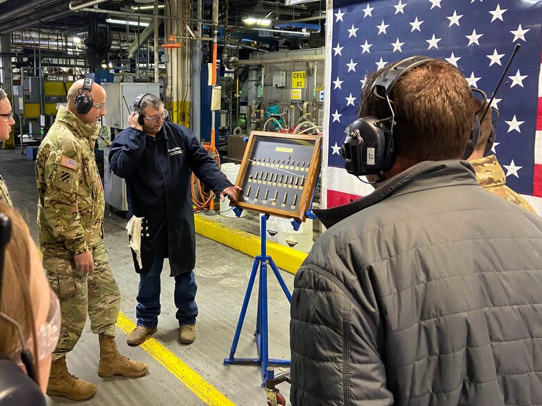 Multiple people, one of which is in a military uniform, look at ammunition rounds with an American flag in the back ground.