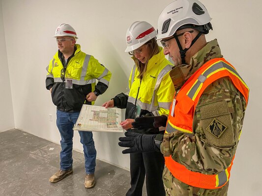 A man in a military uniform, a construction vest and hard hat speak with a woman in a construction jacket and hard hat, while another person watches from near by.