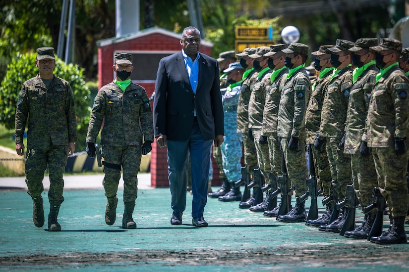 Two men in military attire and a third in business attire walk past a line of soldiers.