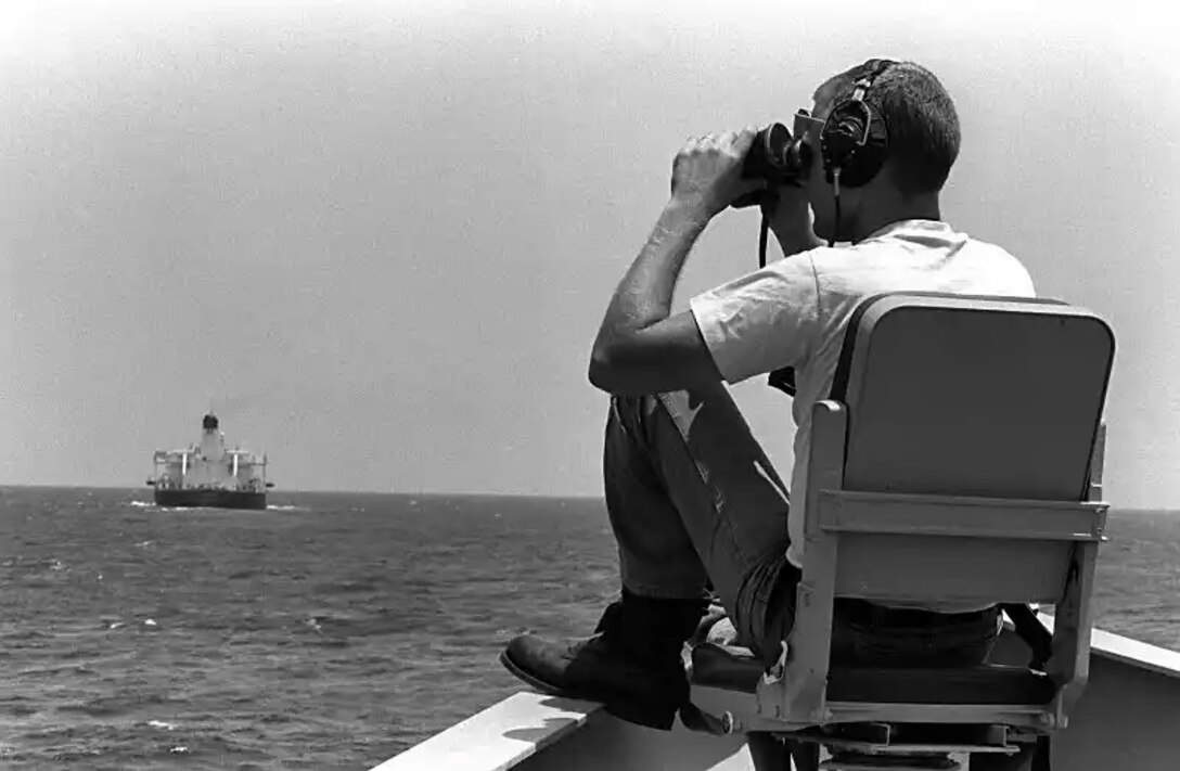 A man sits outside on a chair with his legs up on the ships rail and binoculars up to his eyes as he scans for threats. behind him in he distance is an oil tanker his ship is guarding.