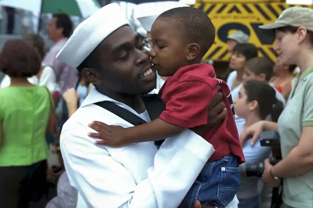 A man in his Navy dress white uniform holds his small son up and hugs him. Many people are in the background.