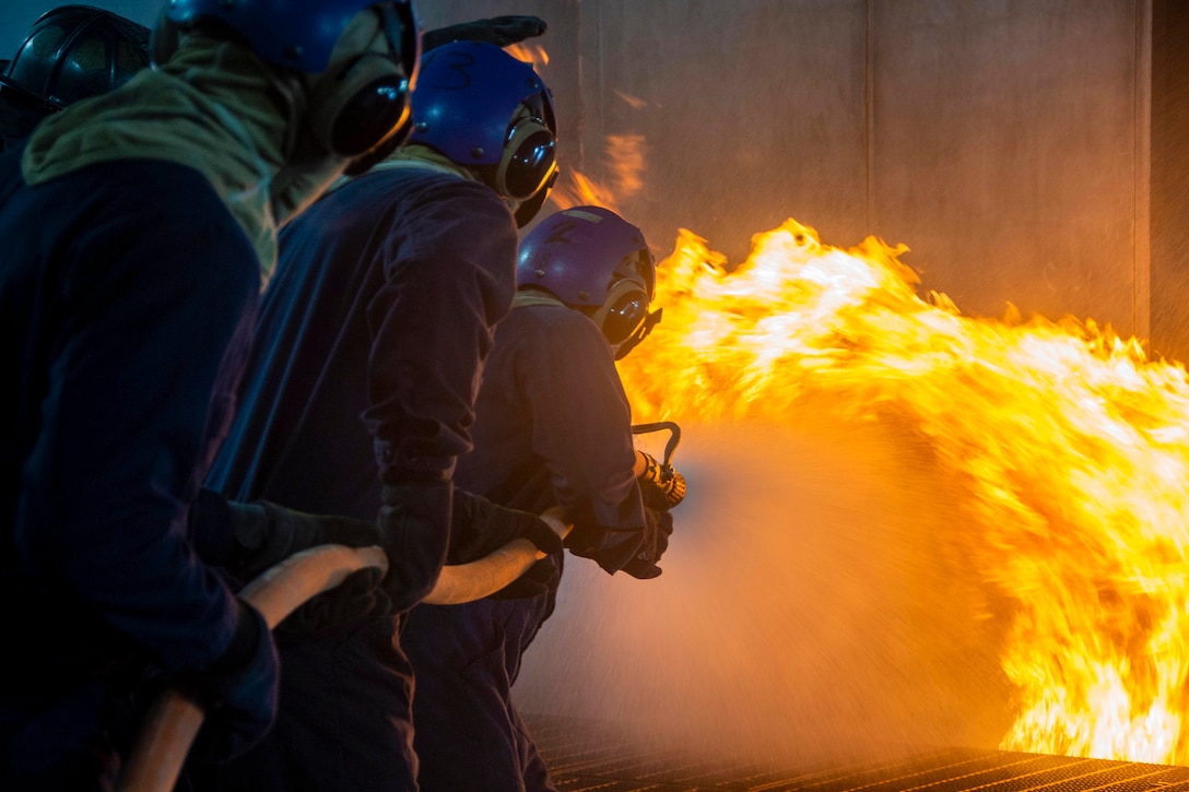Sailors hold onto a water hose as water sprays on a fire.