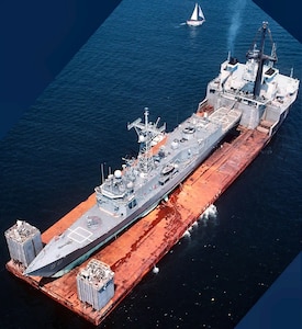 A gray Navy frigate sits out of the water on the orange deck of a heavy lift ship that is underway on a dark blue sea.